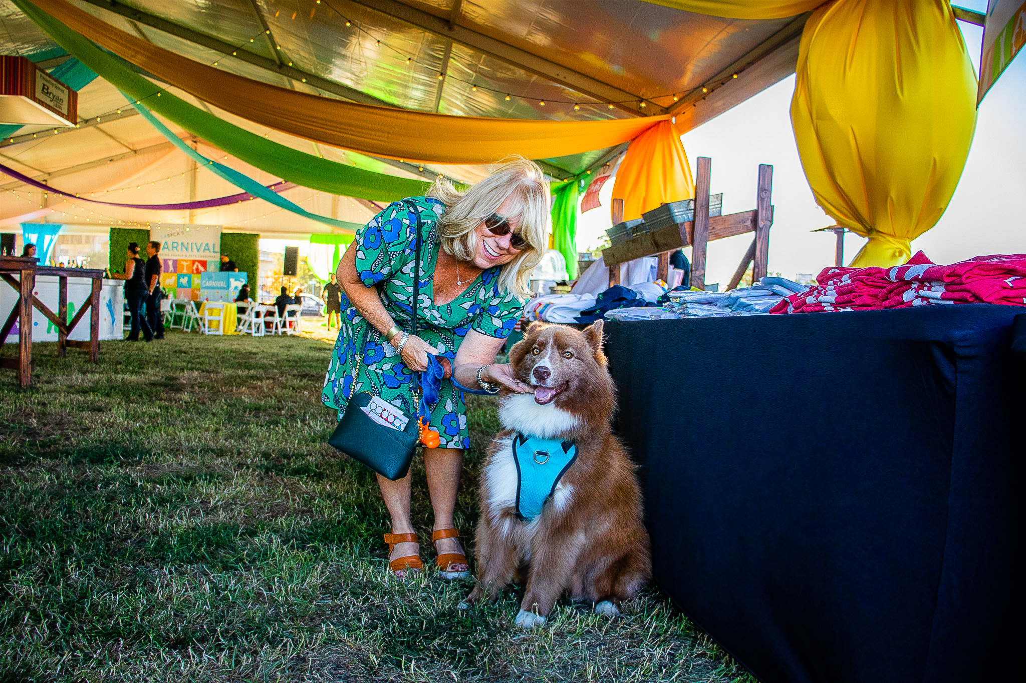 A middle-aged woman with blonde hair and colorful clothes poses for a photo with her fluffy brown and white dog in a Carnival tent