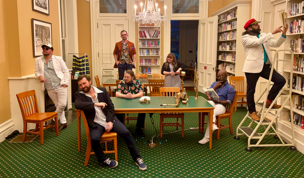 The Where Y'acht band poses for a photo with their 6 male members and single female member in a library to showcase their fun side. 