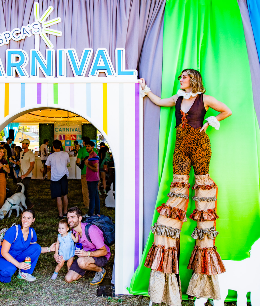 A Yellow Box Circus member, female and on stilts, poses next to the Carnival tent entrance. 